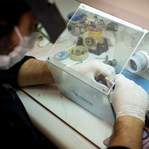 Person working on a small electronic component under a lighted magnifying glass with a visible brand name.