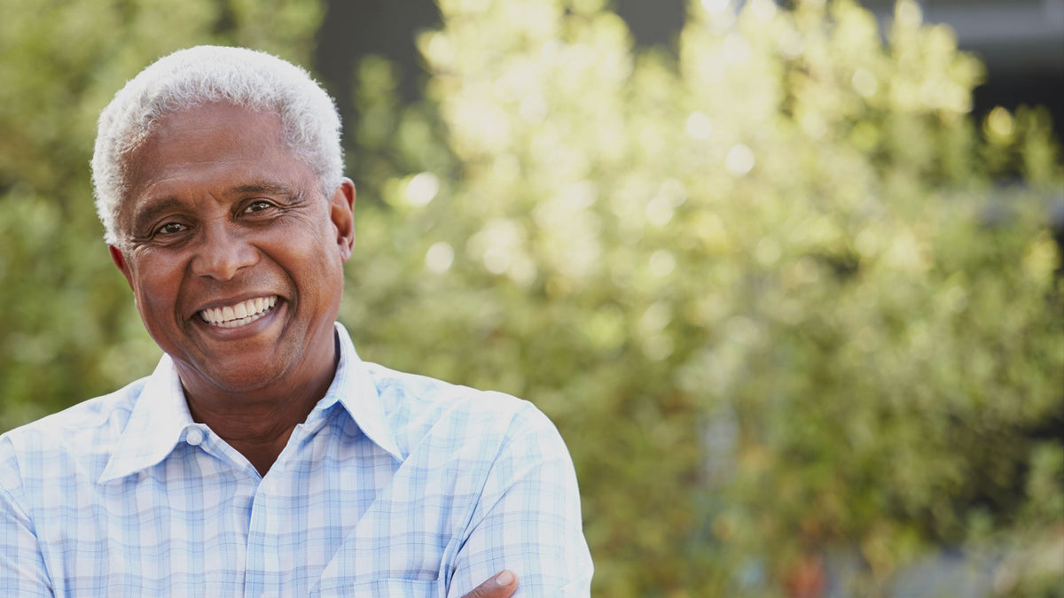 Man with white hair wearing a light blue plaid shirt standing outdoors with greenery in the background
