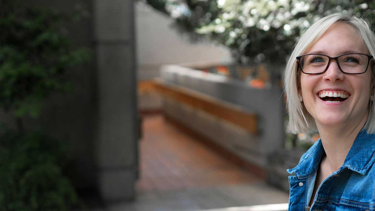 A smiling blonde woman with glasses and a denim jacket, shown in a close-up portrait against a blurred outdoor background.