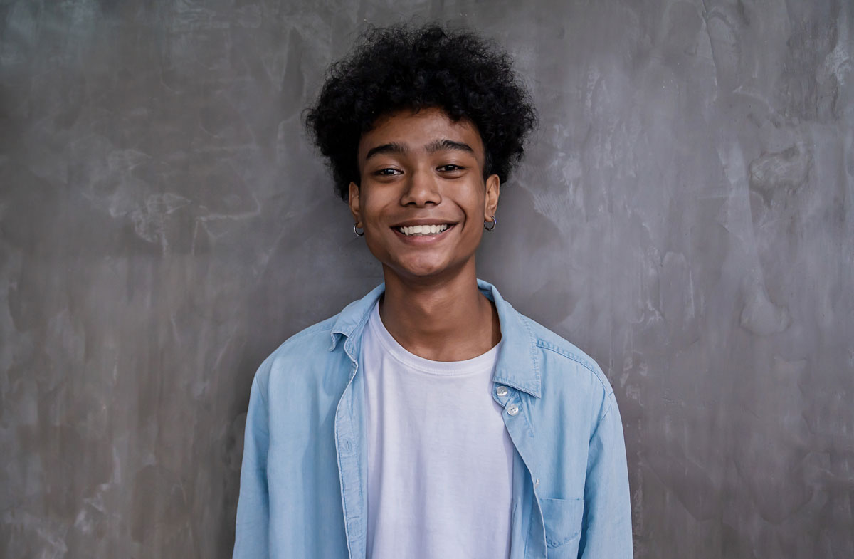 Smiling young man with curly hair wearing a light blue shirt over a white t-shirt, standing against a gray textured wall