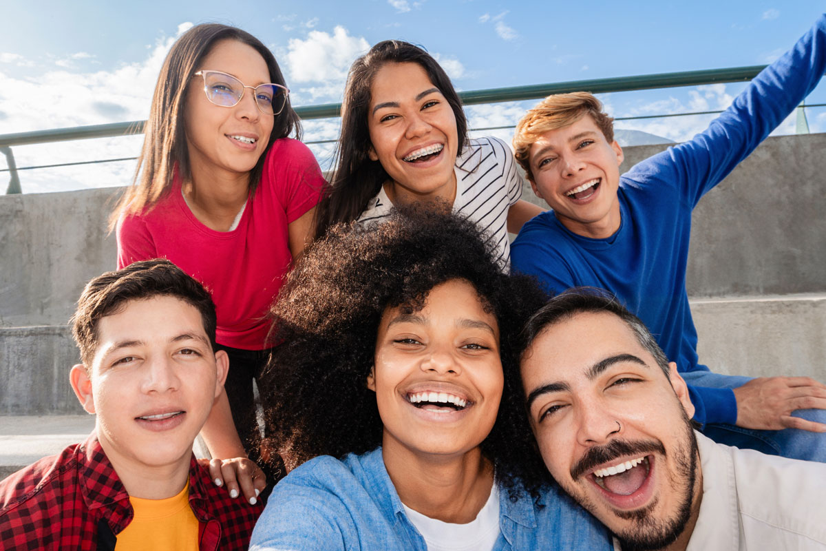a diverse group of six young adults sits outdoors, smiling and taking a cheerful selfie together on a sunny day