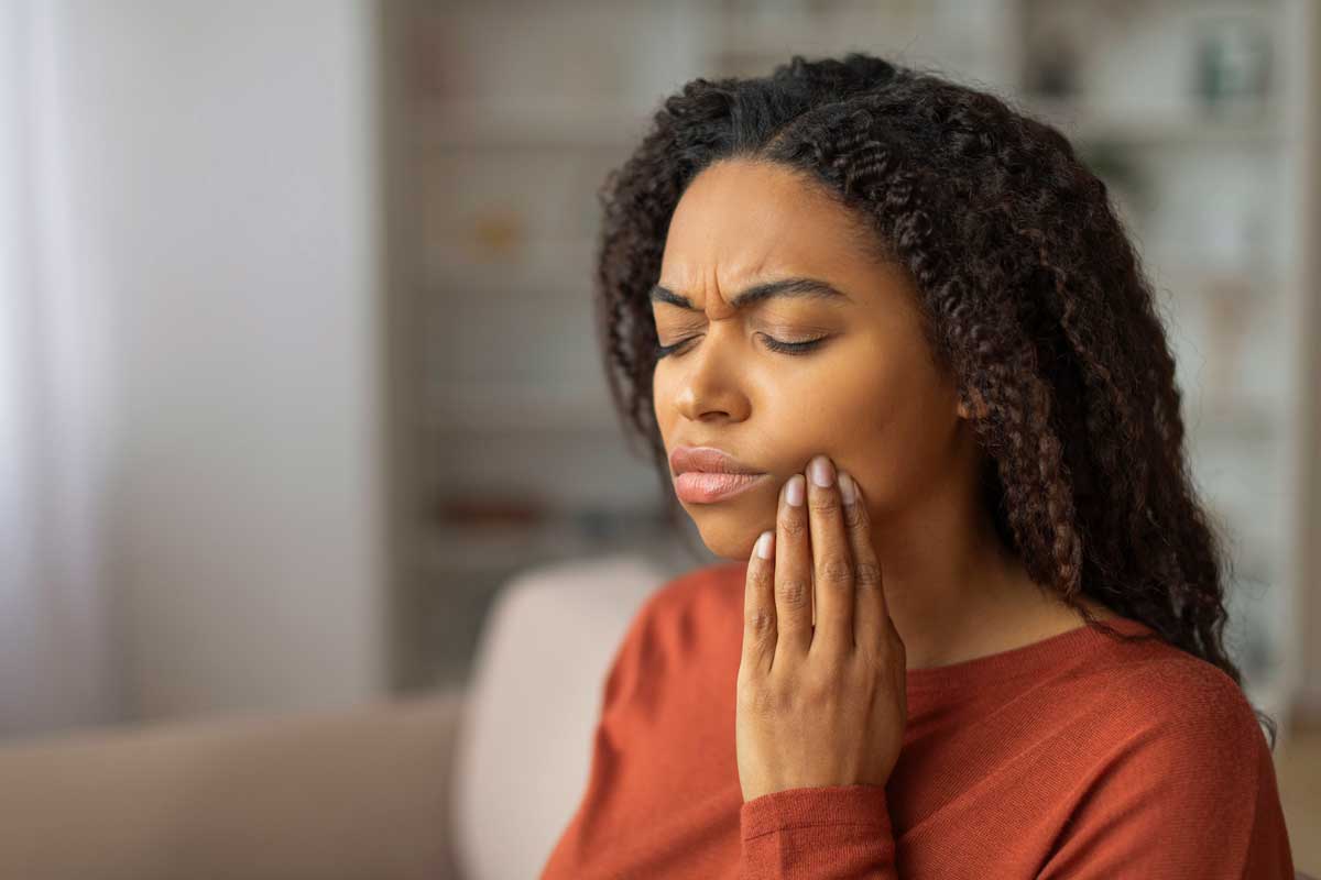 Young woman with curly hair holding her cheek in pain, eyes closed and brows furrowed, indicating jaw discomfort while sitting indoors