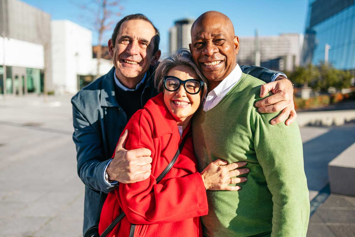 Three older adults smiling and embracing outdoors in a city setting, dressed warmly and enjoying a sunny day together