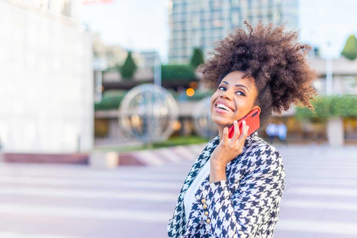 Smiling woman with curly hair holding a red phone outdoors, wearing a houndstooth-patterned blazer