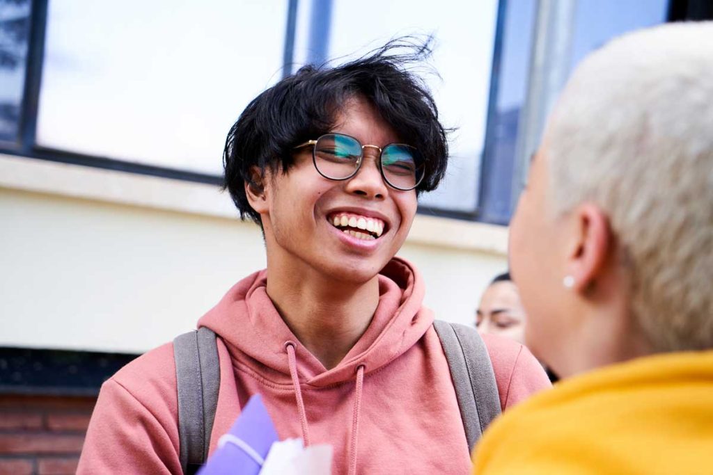 Young man wearing glasses and a pink hoodie smiling and talking with a friend, standing outdoors near a building