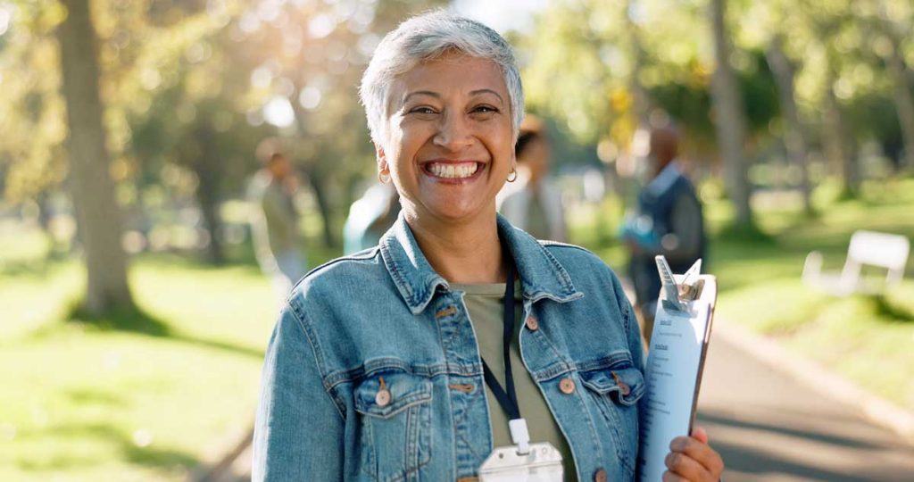 Smiling woman with short gray hair holding a clipboard, standing outdoors in a park with sunlight and people in the background