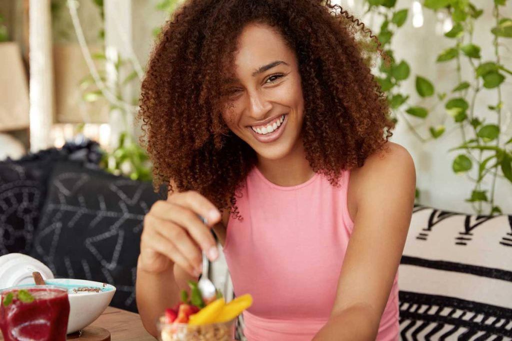 A joyful young woman with curly hair eating a healthy breakfast in a cozy, plant-filled environment