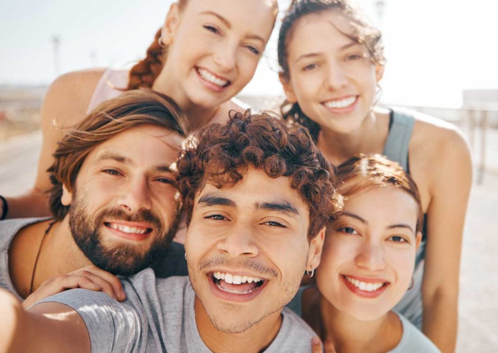 a joyful group selfie of five young friends outdoors, with sunny skies in the background, all smiling broadly at the camer