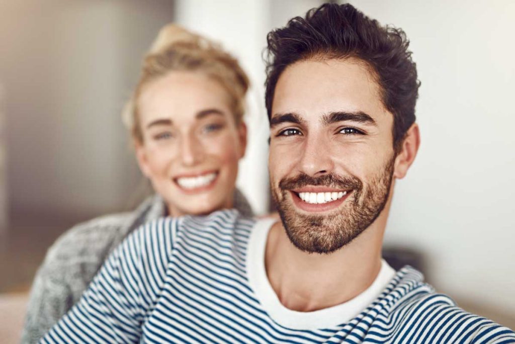 A happy young couple in striped tops, smiling and posing for a selfie, with the focus on the bearded man in the foreground