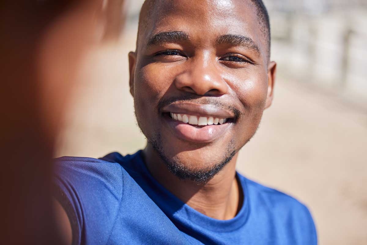 Close-up of a smiling young Black man taking a selfie, wearing a blue shirt with a sunlit beach background