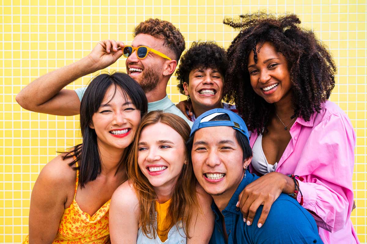 Group of six diverse friends laughing and posing together against a yellow tile background. They are wearing colorful summer clothing and sunglasses, radiating happiness and camaraderie