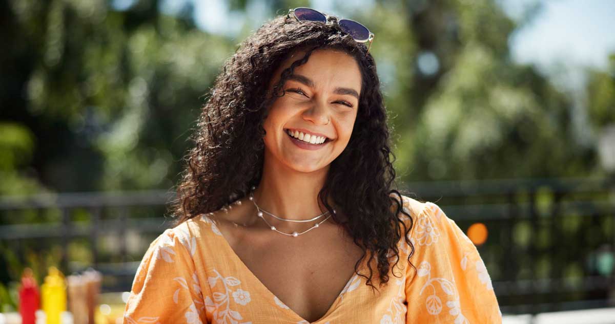 Joyful young woman with curly hair, smiling broadly, wearing a bright orange dress and sunglasses on her head in a sunny, outdoor setting with lush greenery in the background