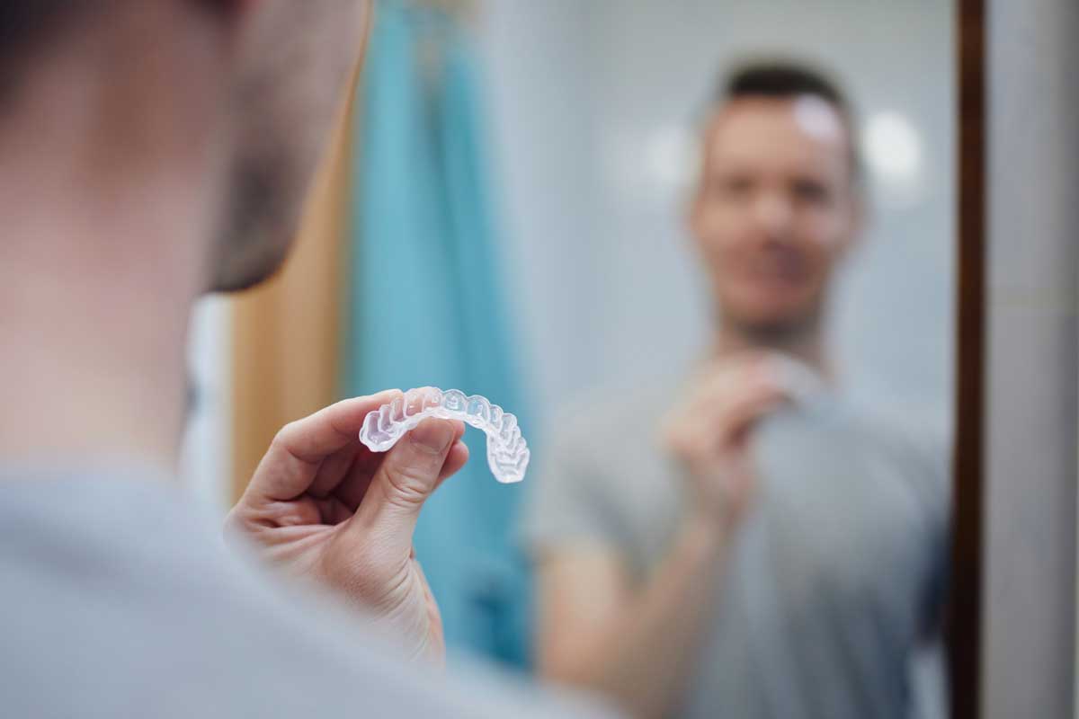 man holding a clear nightguard in focus with his reflection blurred in the bathroom mirror, preparing for dental care.