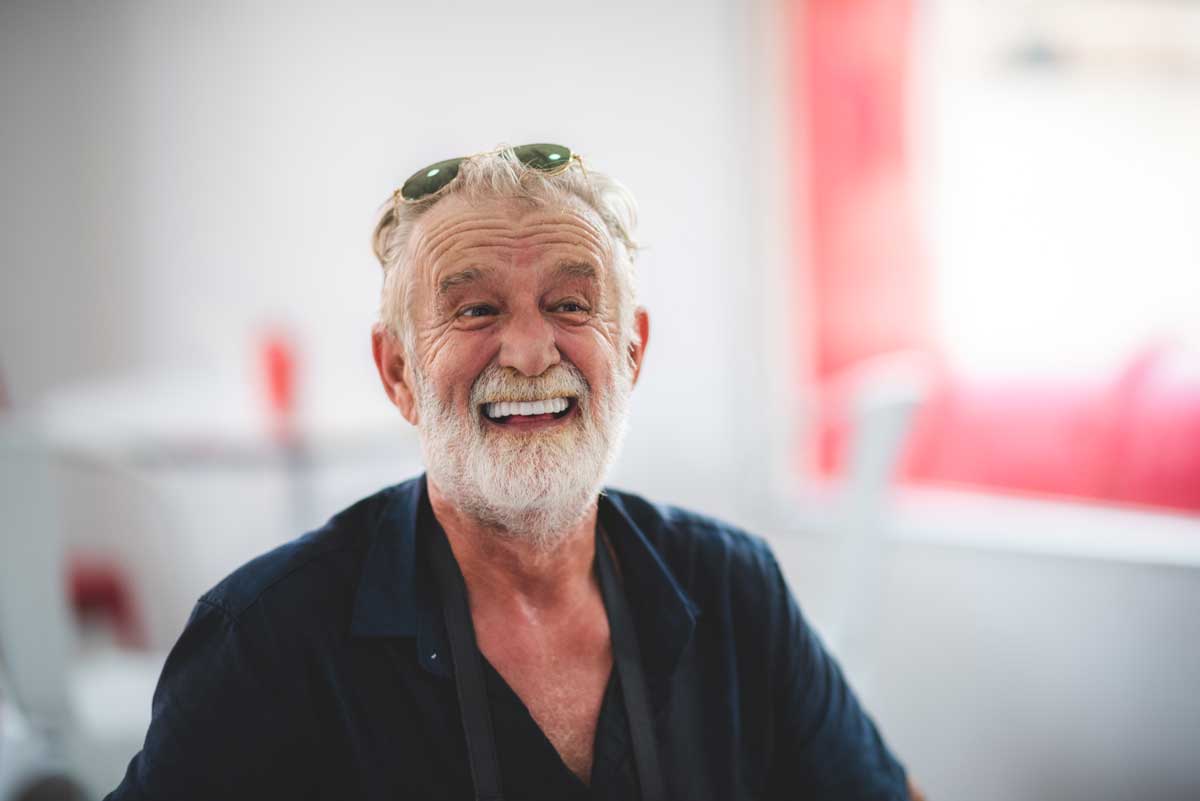 portrait of a cheerful elderly Caucasian man with a white beard, smiling broadly, wearing sunglasses on his head and a dark shirt in a brightly lit room