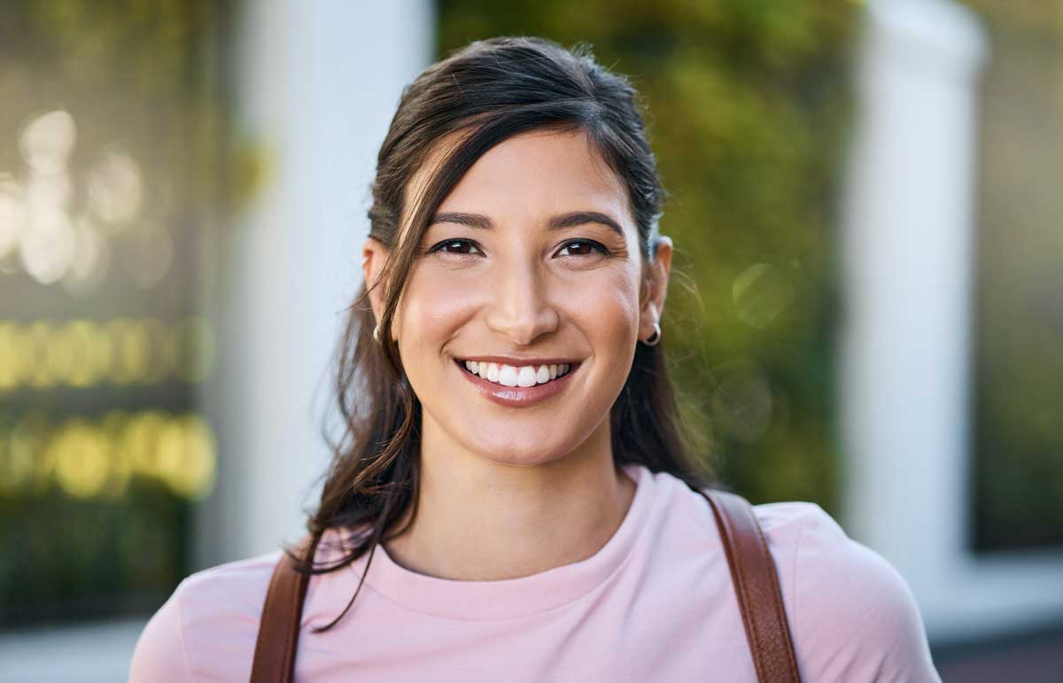 portrait of a smiling young Latina woman wearing a pink shirt outdoors with soft, blurred greenery in the background