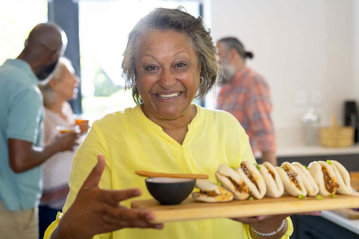 Smiling woman in a yellow shirt holding a tray of bao buns at a casual indoor gathering with friends in the background