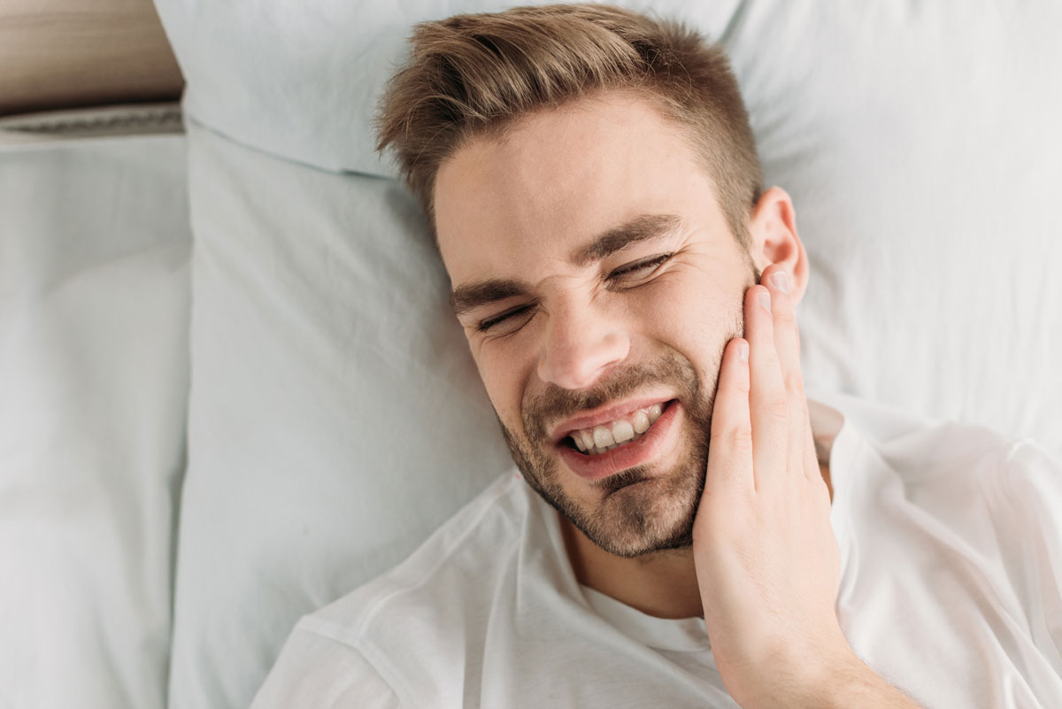 Man lying in bed with jaw pain, holding his face—illustrating symptoms of TMJ disorder and raising the question: do nightguards help TMJ?