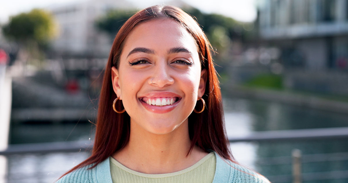 Young woman smiling outdoors near a waterfront, wearing hoop earrings and a light green sweater