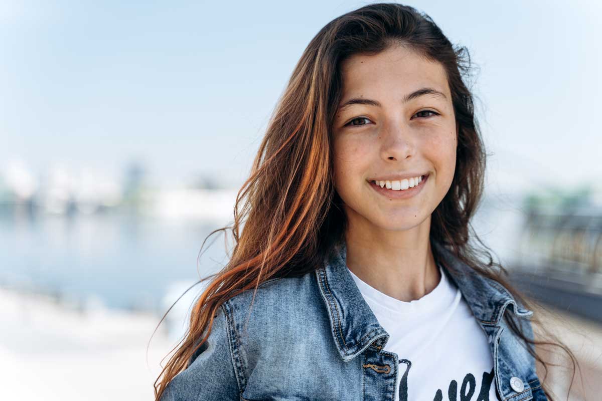 Confident teen girl smiling outdoors in denim jacket near waterfront