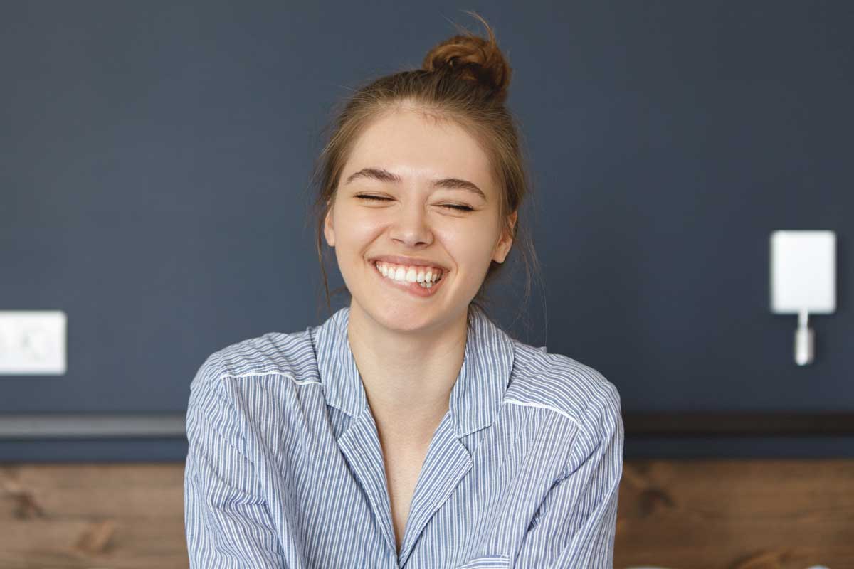 Young woman with hair in a bun smiling with eyes closed, wearing striped pajamas and sitting in a cozy, modern room