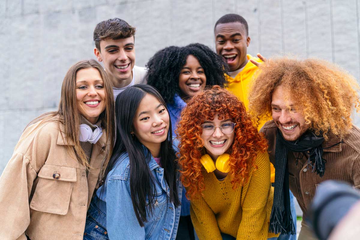 Group of diverse young adults smiling and posing for a photo outdoors, showcasing joyful expressions and casual, colorful outfits.