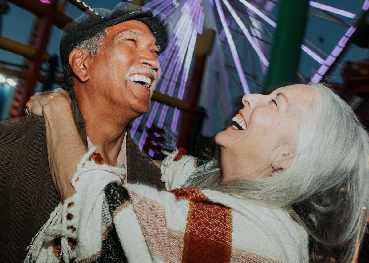 Joyful older couple laughing together at a nighttime amusement park, with a glowing Ferris wheel in the background