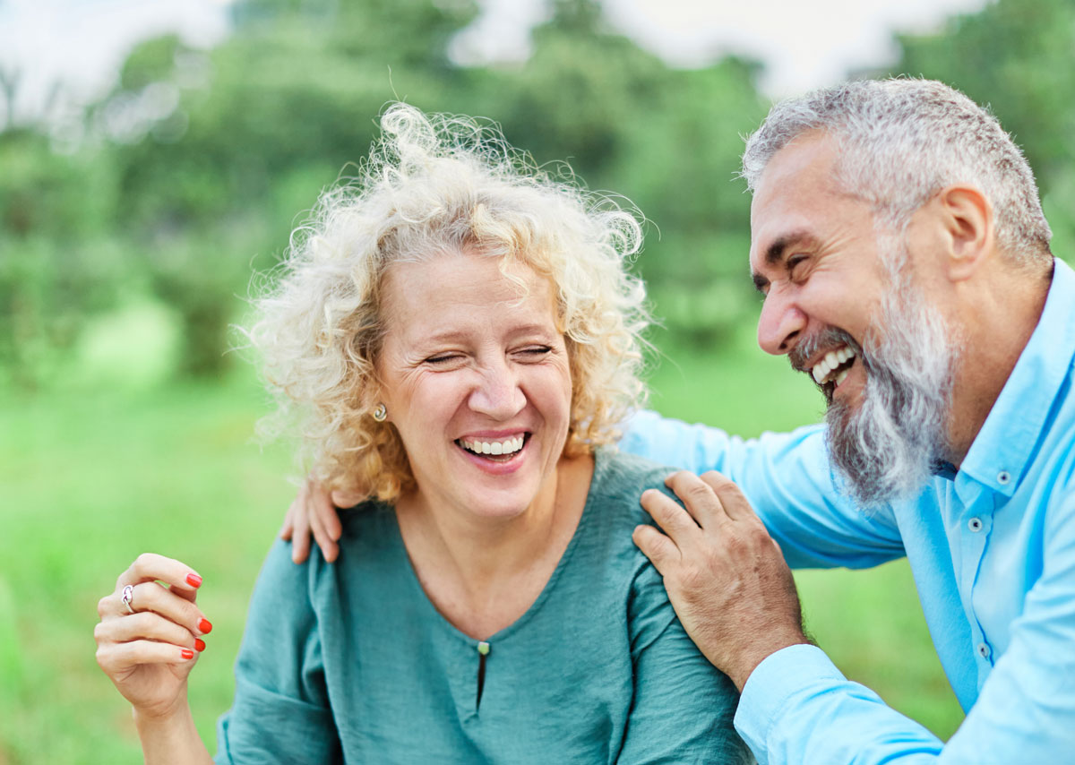 Older couple laughing together outdoors, with the man resting his hand on the woman’s shoulder as they share a joyful moment