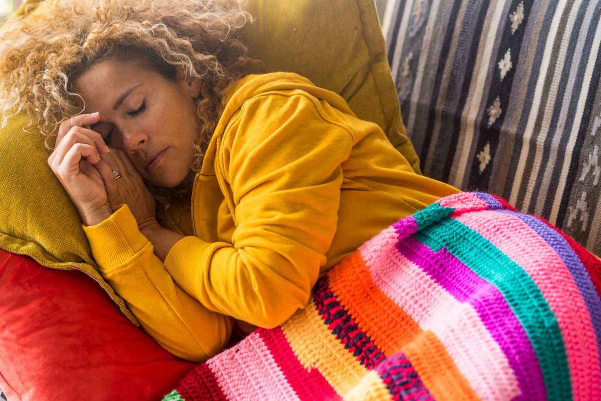 Woman in a yellow hoodie sleeping on a couch, resting her head on a yellow pillow, covered with a colorful striped crochet blanket.