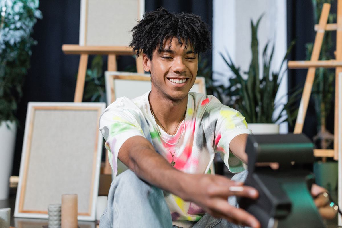 Young man in a tie-dye shirt smiling while sitting indoors with art canvases in the background