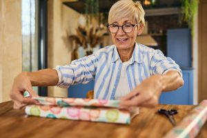 Older woman smiling while wrapping a gift at a wooden table in a cozy home setting.