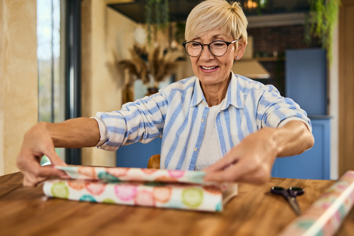 Older woman smiling while wrapping a gift at a wooden table in a cozy home setting.