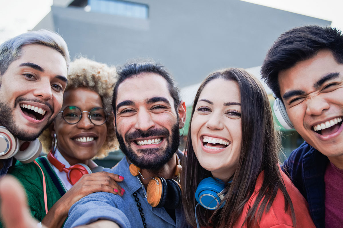 Group of five friends smiling and taking a selfie outdoors, wearing headphones around their necks