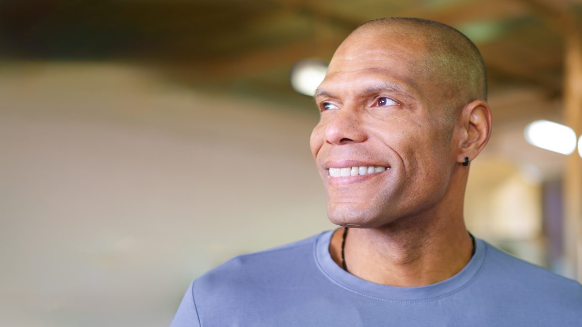 Smiling man wearing a light blue shirt and earring, looking off to the side with soft indoor lighting in the background