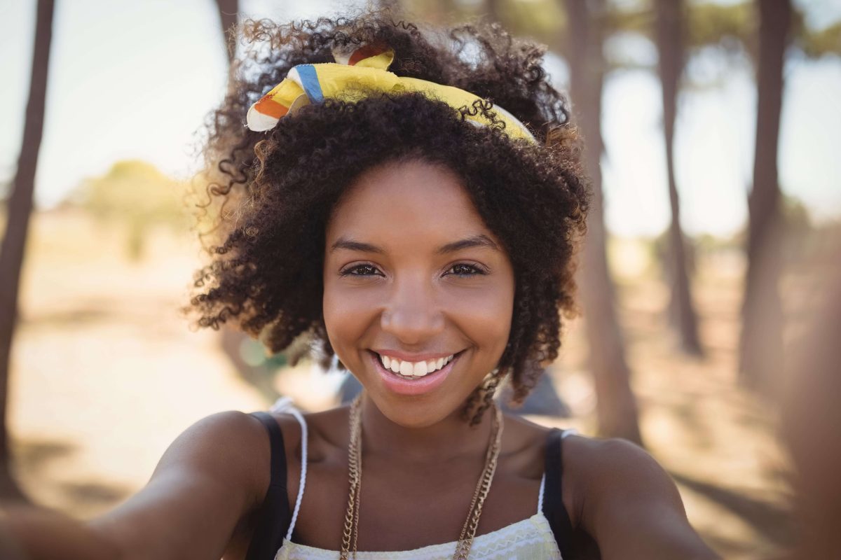 Smiling woman taking a selfie outdoors, wearing a yellow headband and light summer outfit with trees in the background