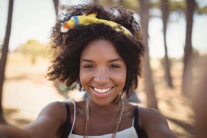 Smiling woman taking a selfie outdoors, wearing a yellow headband and light summer outfit with trees in the background